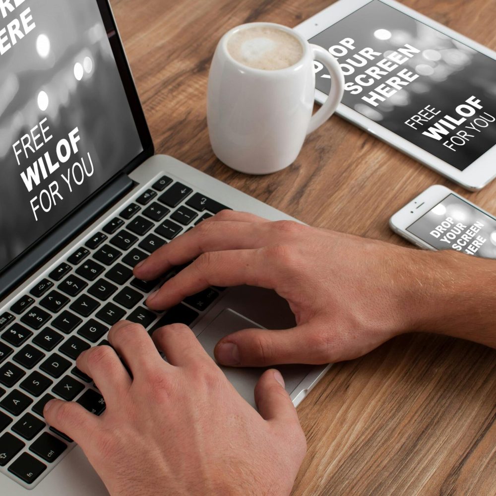 Person using a laptop surrounded by a tablet and smartphone on a wooden desk, displaying sample text.